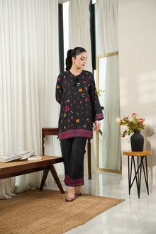 Woman in a black outfit with colorful patterns standing in a room with white curtains and a wooden table.