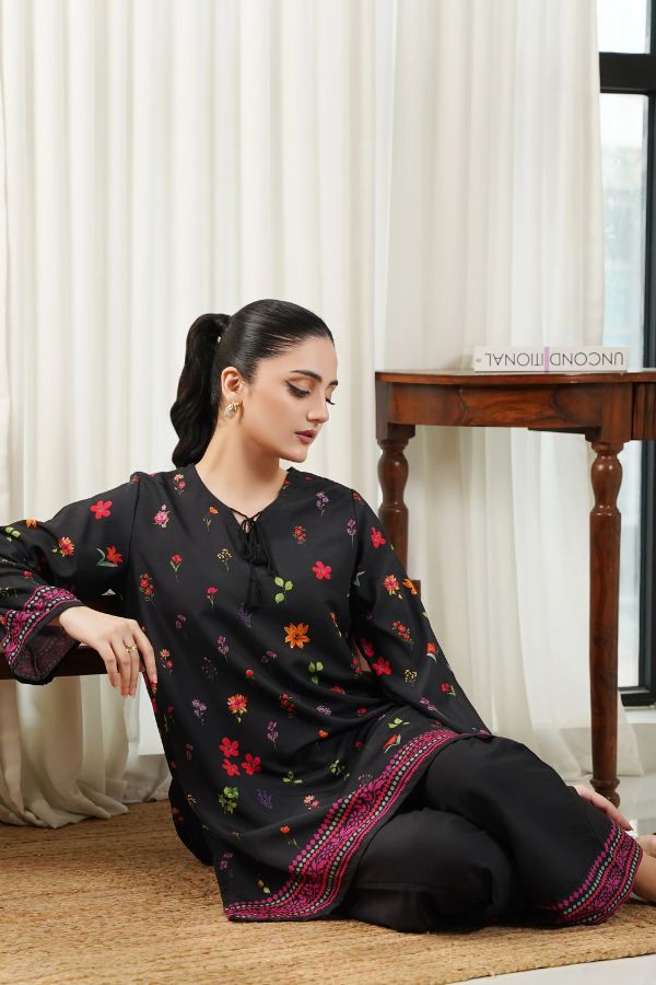 Woman sitting on a wooden floor wearing a black embroidered traditional outfit with colorful patterns.