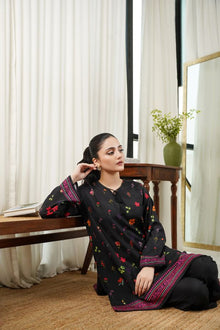 Woman sitting indoors wearing a black traditional outfit with colorful patterns, surrounded by white curtains.