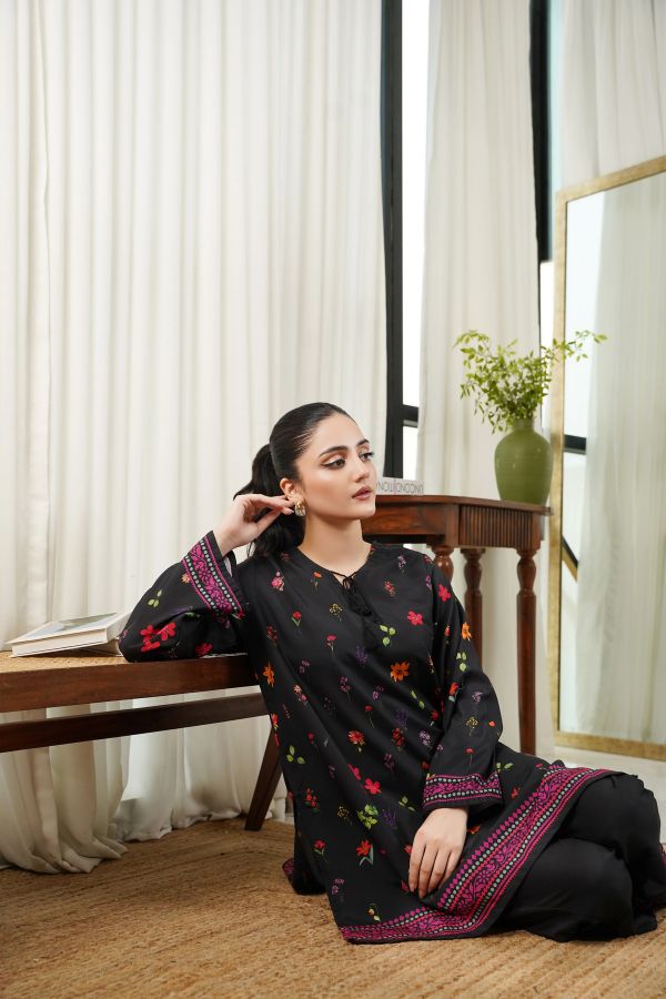 Woman sitting indoors wearing a black traditional outfit with colorful patterns, surrounded by white curtains.