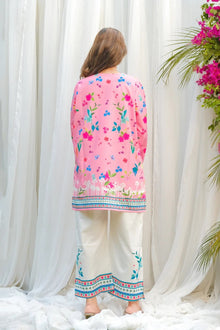 Person wearing a pink floral top and white pants with embroidery, standing against a white curtain.