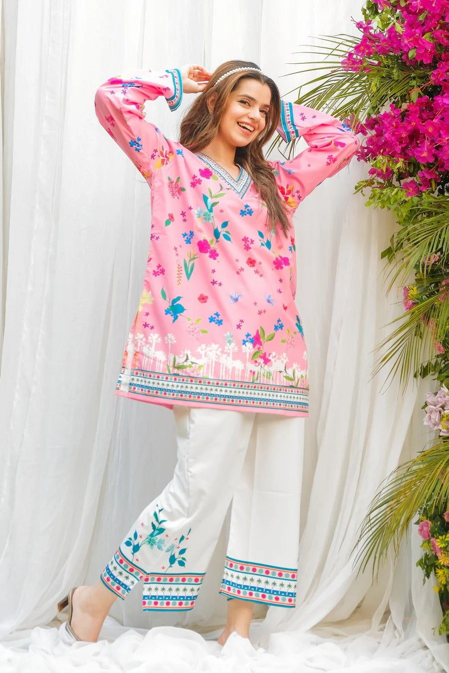 Woman wearing a pink floral top and white pants with decorative patterns, standing against a white curtain with floral decorations.
