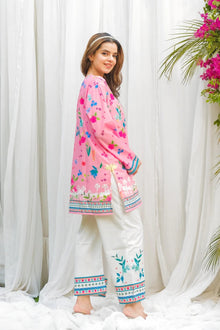 Woman wearing a pink embroidered top and white pants with floral decorations against a white curtain background.