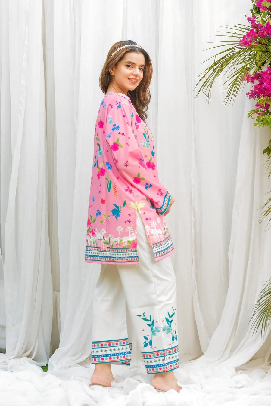 Woman wearing a pink embroidered top and white pants with floral decorations against a white curtain background.