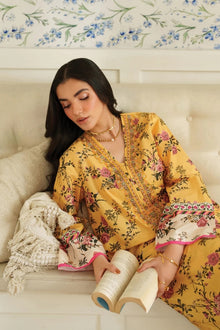 Woman in a yellow floral dress reading a book on a couch with floral wallpaper.
