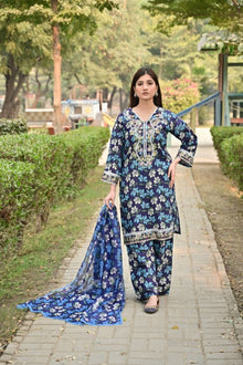 Woman in a blue floral traditional outfit standing on a pathway with greenery.