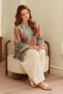 Woman sitting on a chair wearing a colorful embroidered shirt and white pants.
