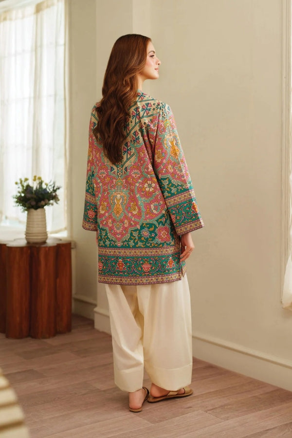 Woman wearing a colorful embroidered top and white pants in a room with a wooden side table and flowers.