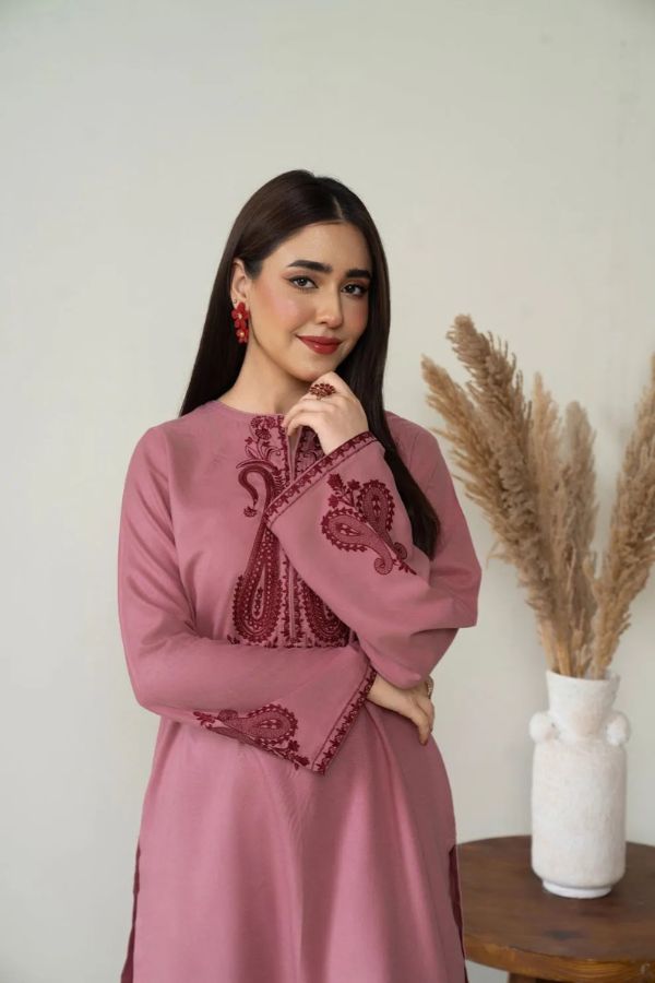 Woman wearing a pink dress with intricate embroidery, standing against a white wall with a vase of dried plants.