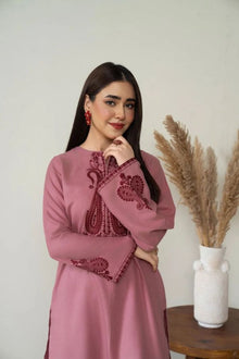 Woman wearing a pink dress with intricate embroidery, standing against a white wall with a vase of dried plants.