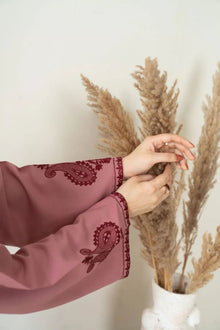 Person wearing a pink long-sleeve garment with intricate embroidery, holding dried plants against a white wall.